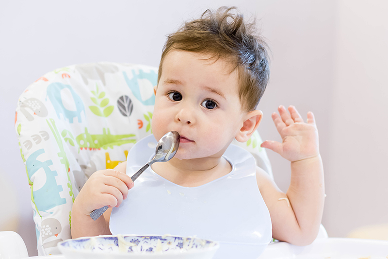 微笑的孩子在厨房吃的食物。1 年岁宝宝吃他们自己的汤。第一次的固体食物，如何教孩子用勺子吃饭。可爱的宝宝，吃午饭用勺子吃饭.jpg
